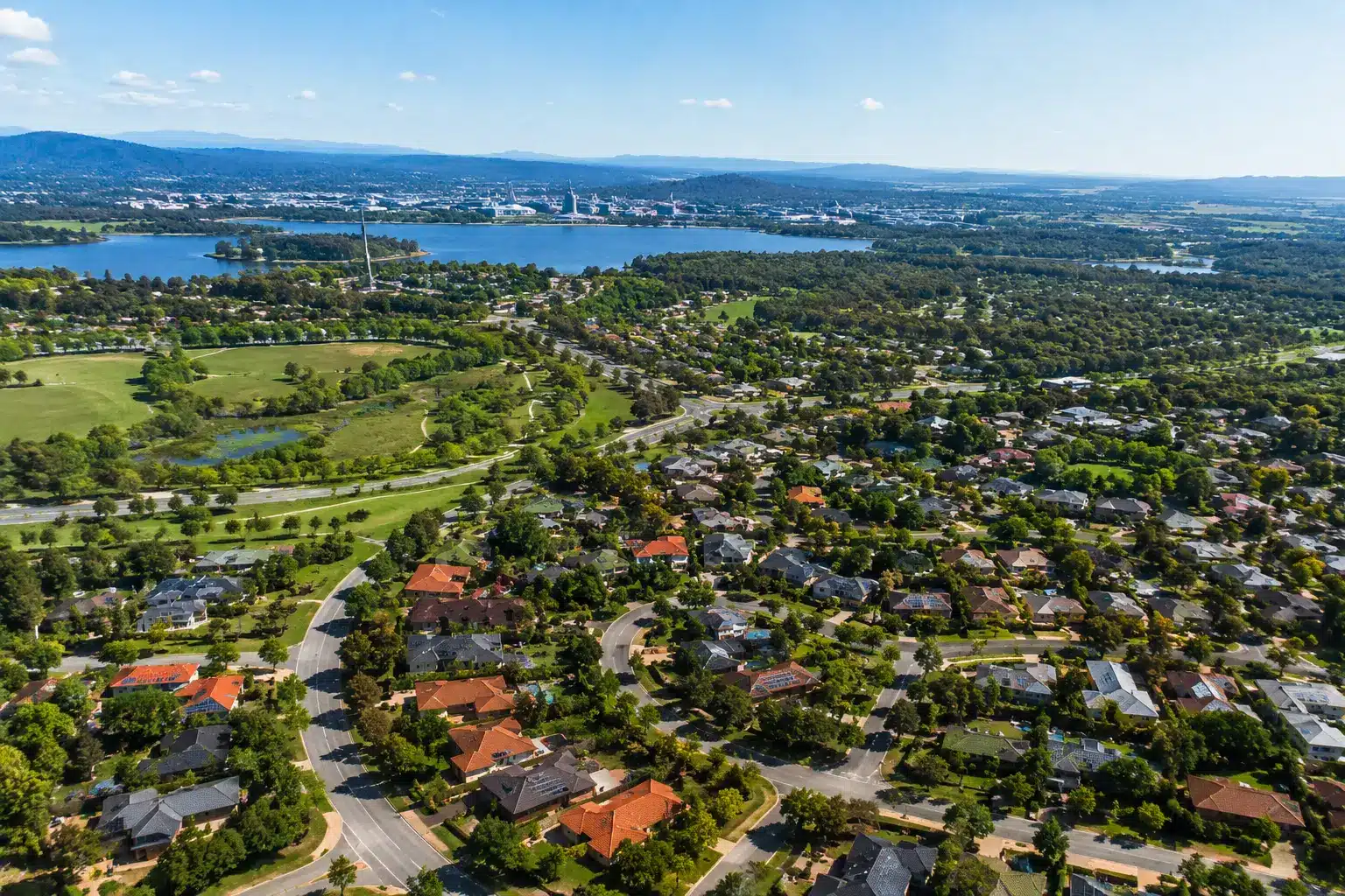 Aerial view of Canberra suburban neighborhoods with residential streets, green parks, and tree-lined roads on a bright sunny day.