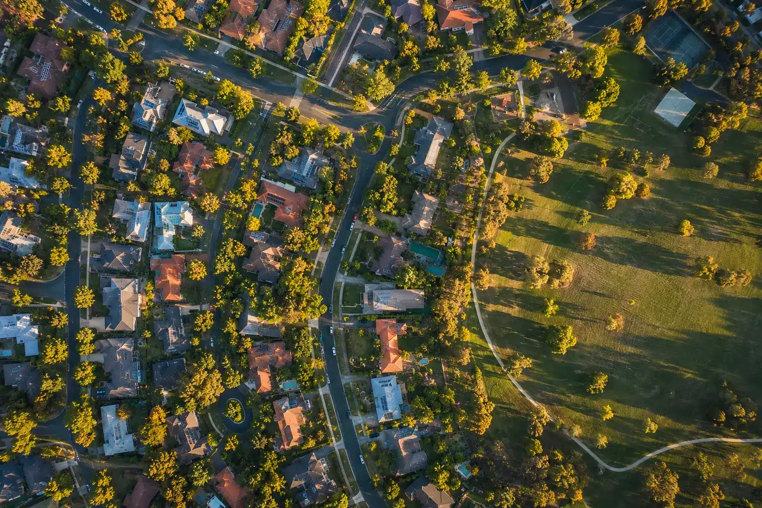 Top-down aerial view of a suburban Canberra neighborhood with tree-lined streets, residential homes, and a large green park during golden hour.